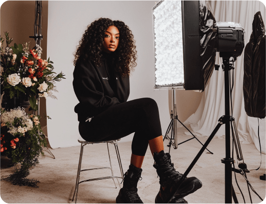 models Fashion model with curly hair seated in a studio setup, surrounded by lighting equipment and floral arrangements.